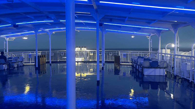 雨で濡れた早朝のデッキに青い照明が残る船上風景